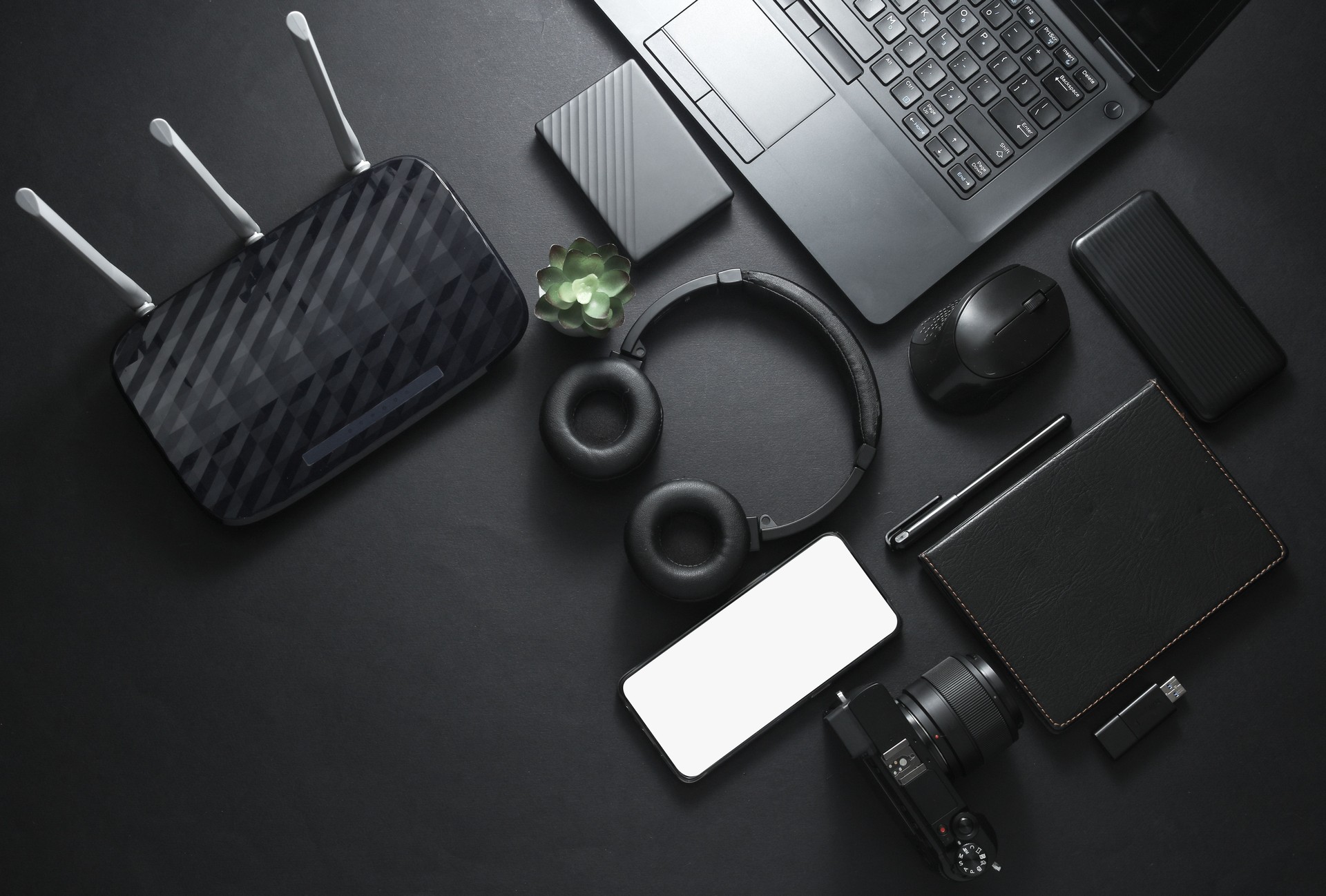 Modern gadgets and accessories in black. Laptop, camera, smartphone, stereo headphones, notepad, external hard drive, USB flash drive on a black background. Photographer Working space. Flat lay. Top view